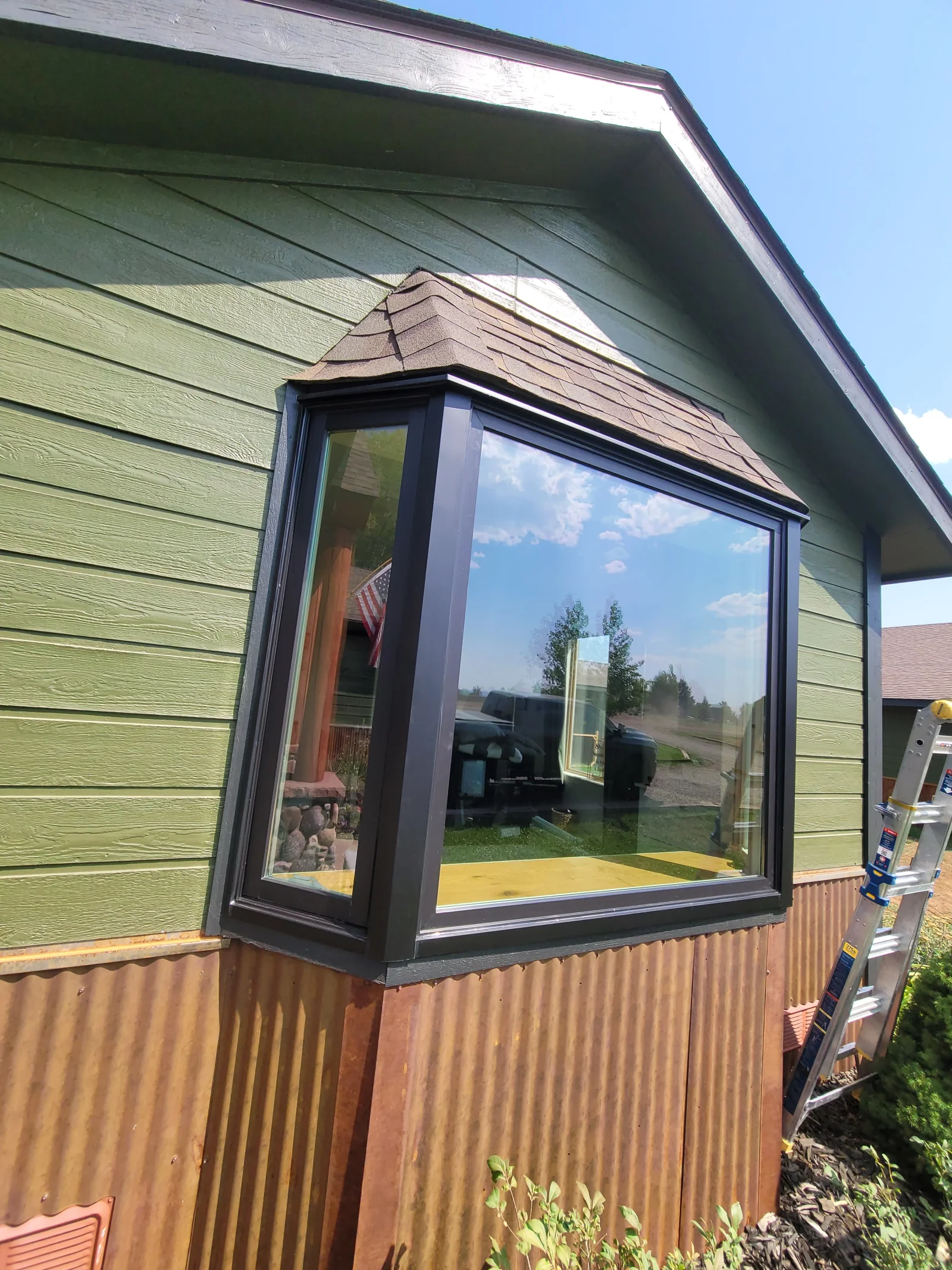 Bay window with dark bronze frames, shingled bay roof, and green lap siding above corrugated metal skirting