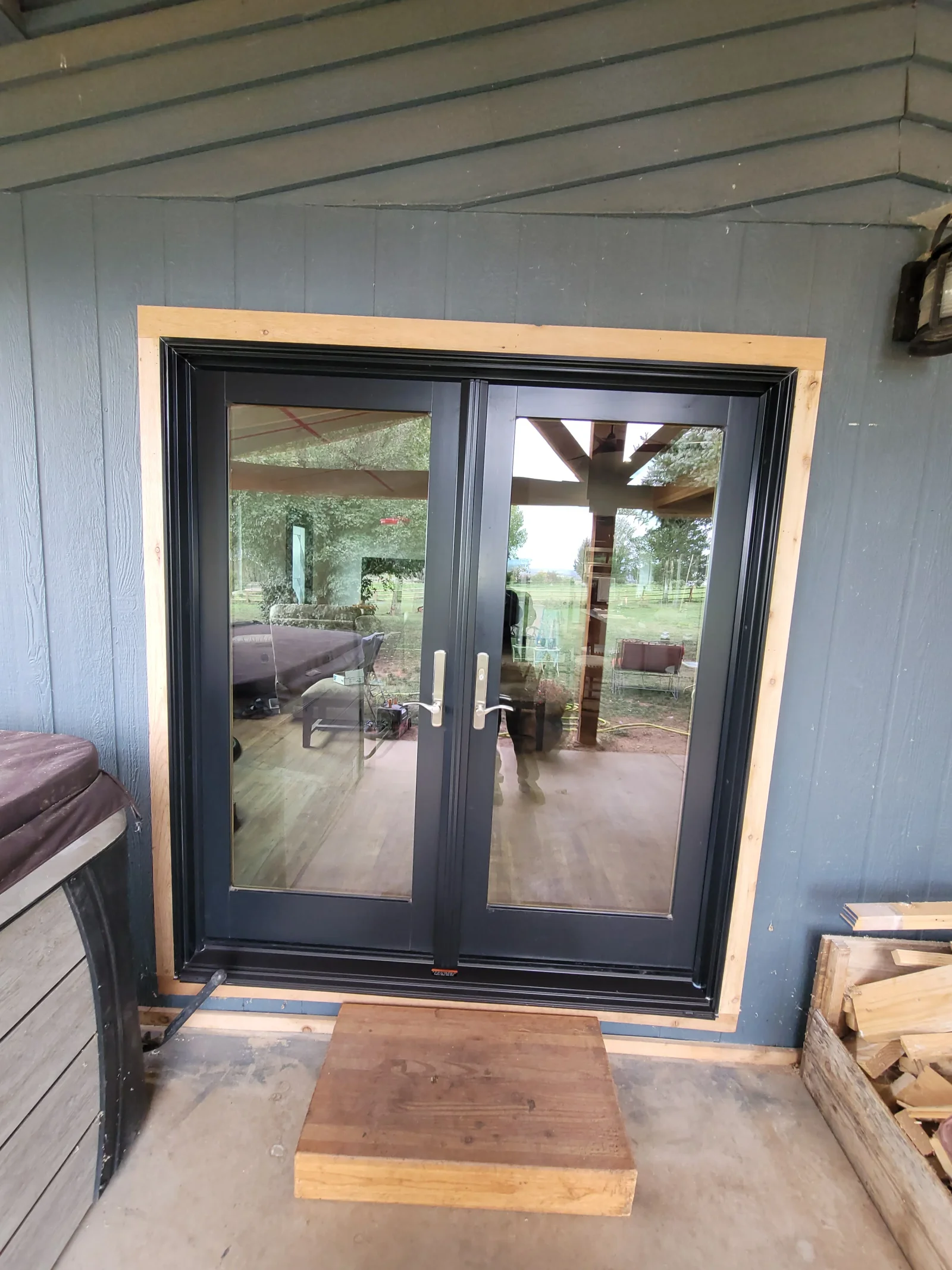 Black double French patio door with full glass panels, silver lever handles, and raw wood trim installed in gray board siding